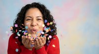 Happy Hispanic woman in red sweater blowing colorful confetti from hands against soft pastel background for celebration and party concepts.