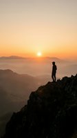 Silhouetted hiker standing on rocky mountain peak at sunrise, gazing over misty valleys and distant mountains bathed in golden light. Perfect for adventure and motivation.