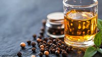 Glass of aged whiskey or bourbon with scattered black and white peppercorns on dark stone background, selective focus on foreground spices and amber liquid.
