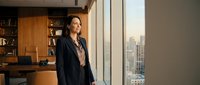 Mature woman in business attire gazing out floor-to-ceiling windows in modern office with city skyline view, wooden desk and bookshelf visible, contemplative professional moment.