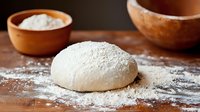 Fresh raw dough ball dusted with flour on wooden surface, ceramic bowl with flour in background, rustic baking preparation in warm lighting.