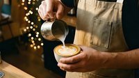 Skilled male barista in apron pouring steamed milk into latte art coffee cup in cozy cafe with festive bokeh lights background.