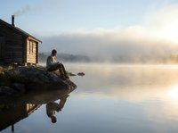 Young woman sitting peacefully by misty lake at sunrise with wooden cabin, perfect reflection in calm water creating serene morning atmosphere.