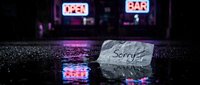 Crumpled paper note with word Sorry lying on wet pavement in rain, glowing neon bar signs reflecting in puddles at night.