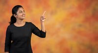 Young South Asian woman in black top pointing up, smiling confidently against vibrant orange background, copy space for text.