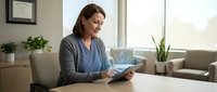 Young woman of mixed ethnicity wearing casual business attire, holding tablet while working at desk in modern office environment with natural light and plants.