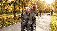 Senior Caucasian man in wheelchair with caring blonde woman in autumn park setting with golden sunlight and fallen leaves creating warm family moment.