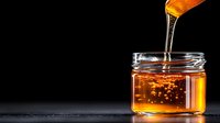 Golden honey dripping from wooden dipper into glass jar against black background, natural sweetener for culinary and wellness applications.