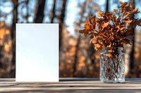 Blank white card mockup with dried oak leaves in vintage glass jar on wooden table outdoors. Autumn forest background creates cozy atmosphere.