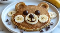 Cute bear-shaped pancake decorated with banana slices, chocolate chips forming eyes and nose, creating playful breakfast for kids on white plate with scattered toppings.