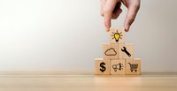 Hand placing wooden cube with lightbulb icon on stack of blocks featuring business symbols including dollar sign, megaphone and shopping cart on desk.