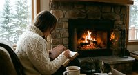 Young woman in cozy cable knit sweater reading book by stone fireplace with warm fire. Relaxing winter indoor scene with coffee cup and peaceful atmosphere.