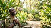 Middle-aged Black man wearing olive green shirt sitting on wooden bench in lush garden setting, hand on chest, contemplative expression, surrounded by green foliage and natural sunlight for wellness and mindfulness concepts.