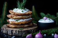 Stack of crispy potato pancakes with sour cream and fresh dill garnish on rustic wooden slice, served with red onions against dark background.