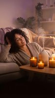 Woman with curly dark hair relaxing on couch surrounded by lit candles and potted plants in warm ambient lighting, ideal for wellness and home relaxation content.