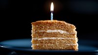 Slice of layered honey cake with cream filling and single burning birthday candle on dark background, dramatic lighting creates festive mood.