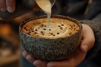 Barista pouring steamed milk into rustic ceramic cup creating latte art pattern, coffee beans scattered on creamy foam surface, cozy coffee shop atmosphere.