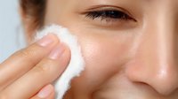 Young Asian woman applying skincare product with cotton pad on cheek, demonstrating facial cleansing routine for healthy glowing skin.