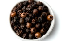 Aromatic black peppercorns in white ceramic bowl on white background, macro view of whole dried spice berries showing texture and natural variations.