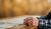 Senior man with age spots and wrinkles wearing plaid shirt rests hand on wooden table in warm natural light, suitable for healthcare, aging, retirement and lifestyle content.