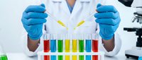 Scientist in white lab coat and blue gloves holding test tubes with yellow liquid above colorful chemical samples for laboratory research and analysis work.
