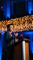 Professional businessman delivering keynote speech at corporate event with warm bokeh lights and blue ambient lighting creating elegant conference atmosphere.