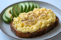 Creamy scrambled eggs on dark bread toast garnished with herbs and black pepper, served with fresh sliced avocado on gray ceramic plate. Close-up food photography.