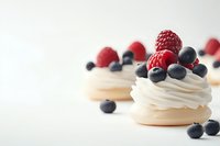 Mini pavlova desserts topped with fresh whipped cream, ripe raspberries and blueberries on white background, selective focus on foreground meringue.