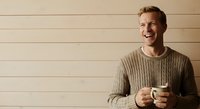 Caucasian male in casual ribbed sweater holding coffee cup against neutral beige wall background, suitable for lifestyle, wellness, and beverage marketing campaigns.