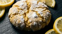 Freshly baked lemon scone with cracked golden surface dusted with powdered sugar, surrounded by bright yellow lemon slices on dark slate background.