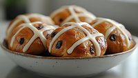 Fresh hot cross buns with glossy glazed tops and raisins served in white ceramic bowl, traditional Easter holiday baked treats with cream cross decoration.