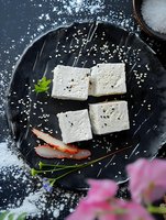 Fresh tofu cubes garnished with sesame seeds and herbs on black plate with sliced radish and pink flowers, dramatic food photography on dark background.