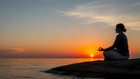 Woman practicing meditation yoga pose on rocky coastline during golden sunset over calm ocean waters creating peaceful mindfulness moment.