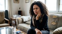 Young African American woman experiencing toothache pain sitting on couch at home holding jaw with hand expressing discomfort in bright living room interior.