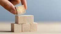 Hand placing wooden block with glowing light bulb icon on top of stacked blocks, representing innovative idea or business solution concept.