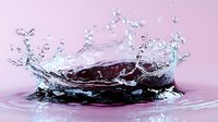 Dynamic water crown splash captured in high speed against pink background, with detailed droplets and ripples creating symmetrical liquid art formation.