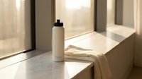 Minimalist bathroom counter with white cosmetic bottle and beige towel near window, showcasing skincare product placement for wellness and beauty marketing.