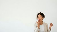 Young woman with curly dark hair and medium skin tone, wearing a beige cardigan over a white top, touching her face thoughtfully while looking upward against a white background.