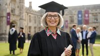 Mature Caucasian woman graduate in cap and gown holding diploma smiling at university campus ceremony with historic stone buildings and fellow graduates.