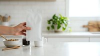 Young woman with medium skin tone pouring coffee into white mug in bright kitchen with potted plant, cereal bowl, and wooden utensils on counter for morning breakfast routine.