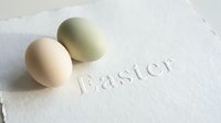 Two pastel Easter eggs in soft beige and sage green rest on textured handmade paper with embossed Easter lettering, minimal spring holiday concept.