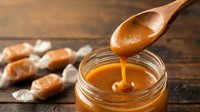Golden caramel sauce dripping from wooden spoon into glass jar with soft candies on rustic wood background for dessert recipes and confectionery.
