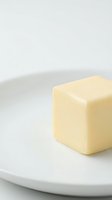 Cube of creamy butter on white ceramic plate against neutral background, suitable for culinary, food styling, cooking, and recipe content.