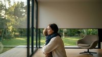 Caucasian mother hugging young child indoors near large window, warm family moment in modern minimalist home with garden view.