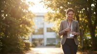Professional businesswoman walking outdoors using smartphone during golden hour commute in urban park setting for corporate lifestyle content.