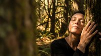 Young woman with closed eyes leaning against tree trunk in peaceful forest setting, connecting with nature during golden hour sunlight filtering through trees.