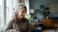 Mature woman with gray hair smiling while looking out window in modern home interior with plants and natural light for lifestyle and wellness content.