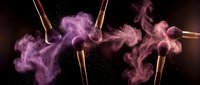 Professional makeup brushes with vibrant purple powder explosion creating dramatic cosmetic dust clouds against dark background for beauty advertising.