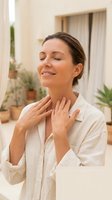 Serene woman applying skincare cream to neck with closed eyes in bright modern home interior with plants for wellness and beauty routine.
