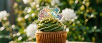 Matcha cupcake with green frosting and a colorful Happy Birthday topper, set against a lush garden background with white flowers.